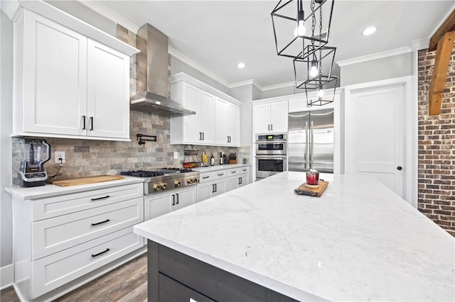 kitchen featuring white cabinetry, wall chimney range hood, appliances with stainless steel finishes, decorative light fixtures, and crown molding