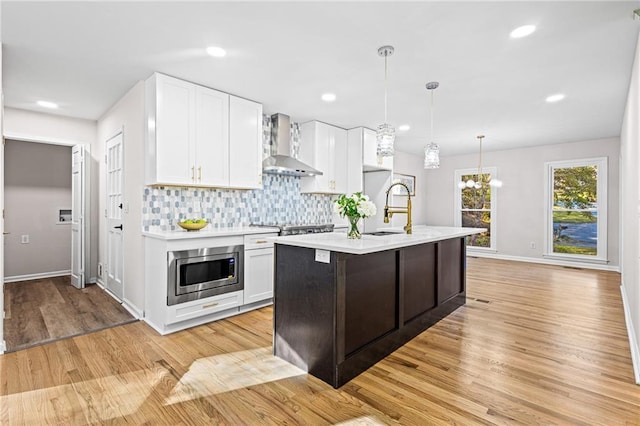 kitchen with white cabinets, stainless steel microwave, light countertops, wall chimney range hood, and a sink