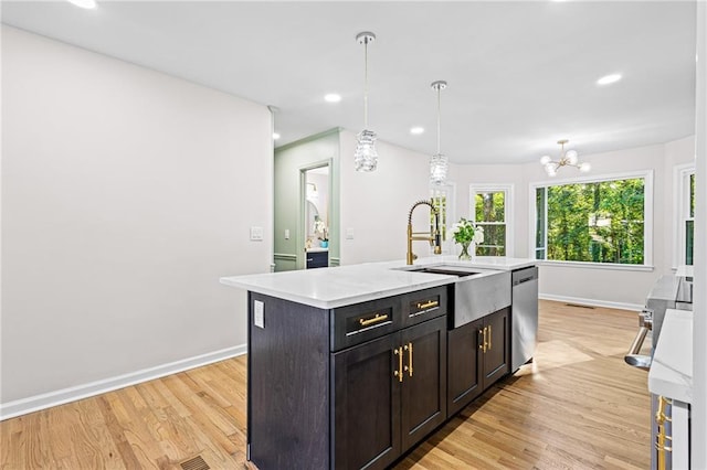 kitchen with light countertops, hanging light fixtures, stainless steel dishwasher, light wood-style floors, and an island with sink