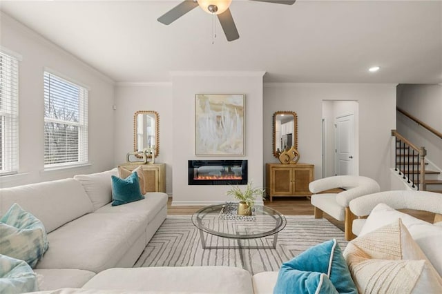 living area with ceiling fan, a glass covered fireplace, ornamental molding, wood finished floors, and stairs