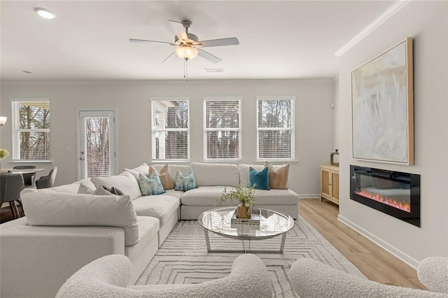 living room with baseboards, a glass covered fireplace, light wood-type flooring, ornamental molding, and a ceiling fan