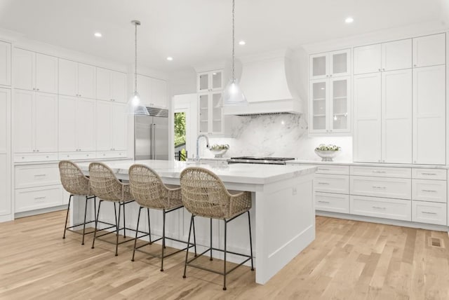 kitchen featuring sink, premium range hood, white cabinetry, stainless steel built in refrigerator, and an island with sink