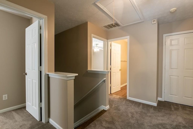 hall featuring attic access, visible vents, dark colored carpet, a textured ceiling, and an upstairs landing