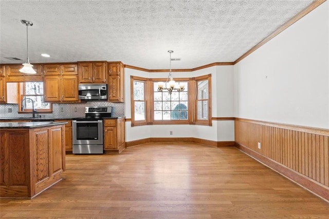 kitchen featuring brown cabinets, a wainscoted wall, stainless steel appliances, dark countertops, and light wood-type flooring