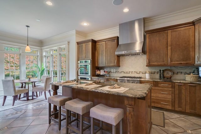 kitchen with an island with sink, pendant lighting, wall chimney range hood, dark stone counters, and backsplash