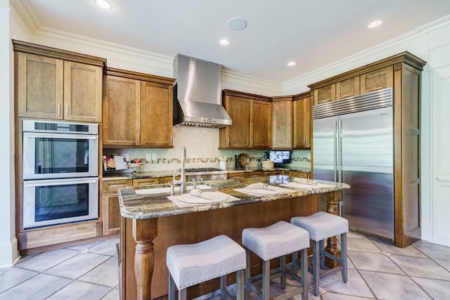 kitchen featuring appliances with stainless steel finishes, dark stone counters, tasteful backsplash, wall chimney range hood, and a center island with sink