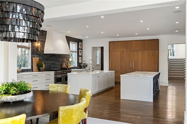 kitchen with a center island with sink, double oven range, dark wood-type flooring, and wall chimney range hood