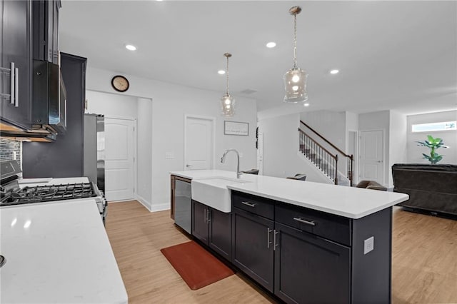 kitchen with light wood-type flooring, stainless steel appliances, sink, decorative light fixtures, and an island with sink