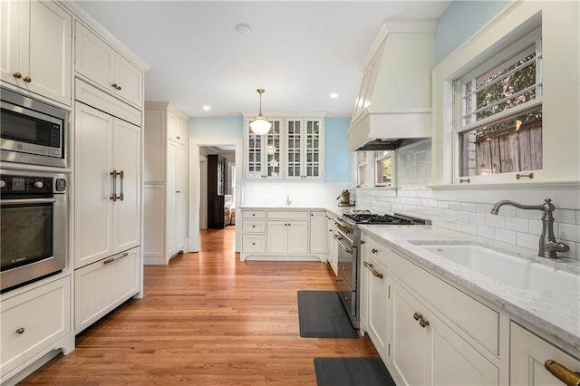 kitchen featuring custom exhaust hood, decorative backsplash, glass insert cabinets, a sink, and built in appliances