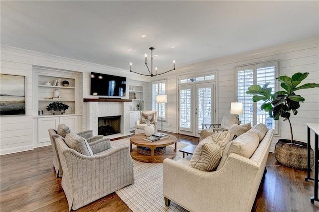 living room featuring a brick fireplace, built in shelves, dark wood-style floors, and a notable chandelier