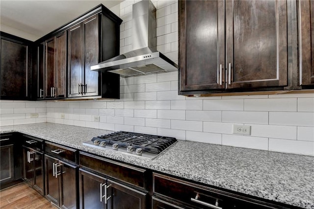 kitchen with light stone countertops, dark brown cabinets, wall chimney range hood, stainless steel gas cooktop, and backsplash