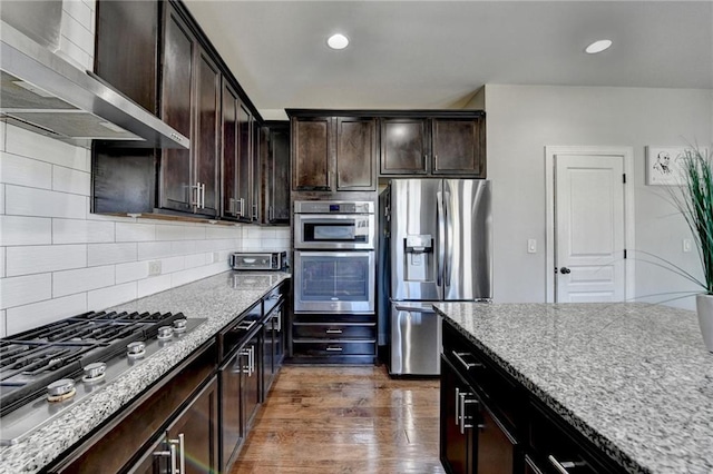 kitchen featuring wall chimney range hood, appliances with stainless steel finishes, dark brown cabinets, decorative backsplash, and light stone counters