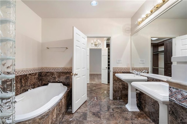 full bath with a relaxing tiled tub, marble finish floor, a wainscoted wall, an inviting chandelier, and two sinks