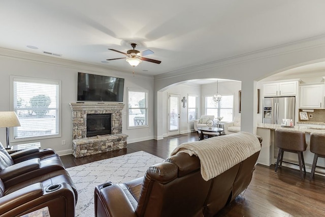 living room with arched walkways, dark wood-style flooring, crown molding, visible vents, and a stone fireplace