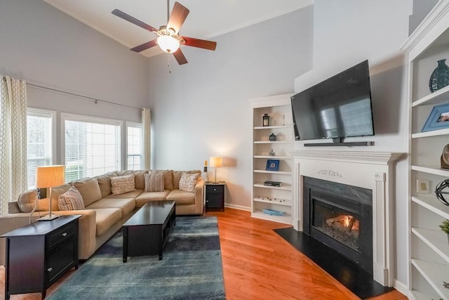 living area featuring light wood-type flooring, a fireplace with flush hearth, a towering ceiling, baseboards, and ceiling fan