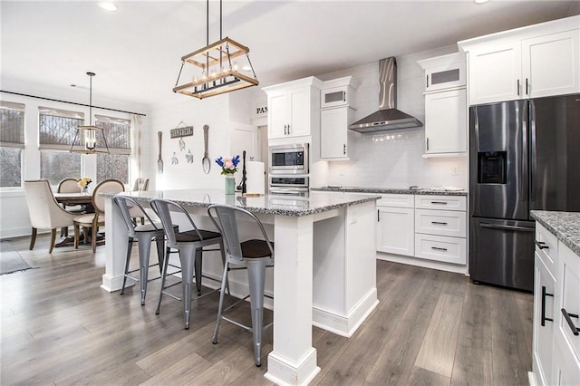 kitchen with wall chimney range hood, a center island with sink, white cabinetry, hanging light fixtures, and stainless steel appliances