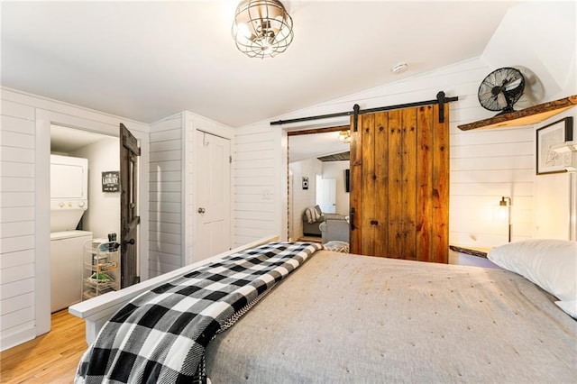 bedroom with a barn door, light wood-style floors, stacked washer / dryer, vaulted ceiling, and wood walls