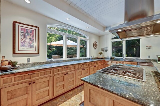 kitchen featuring light brown cabinets, stainless steel gas stovetop, dark stone counters, island range hood, and sink