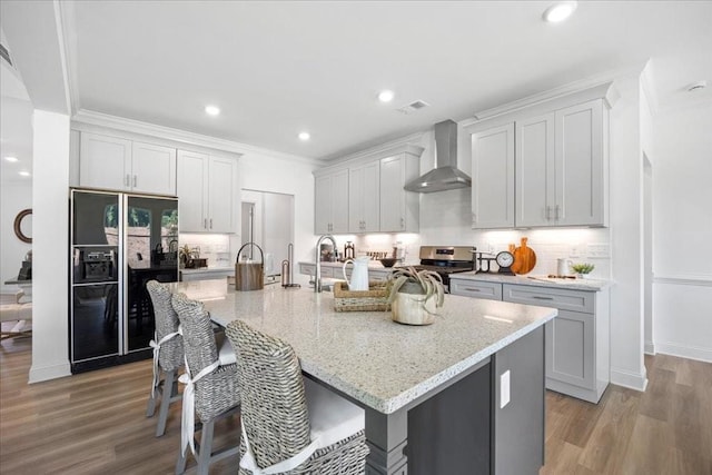 kitchen featuring a kitchen island with sink, black refrigerator with ice dispenser, stainless steel range oven, light stone counters, and wall chimney exhaust hood