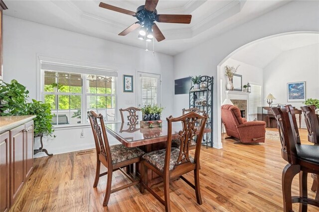 dining space featuring light wood finished floors, arched walkways, a tray ceiling, ceiling fan, and a fireplace