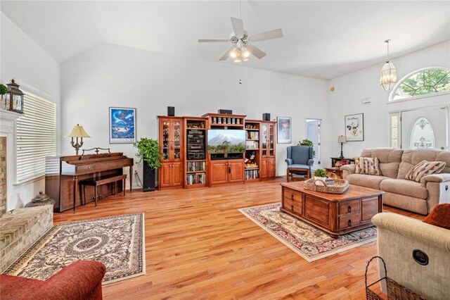 living room featuring high vaulted ceiling, light wood-type flooring, ceiling fan, a chandelier, and a fireplace with raised hearth