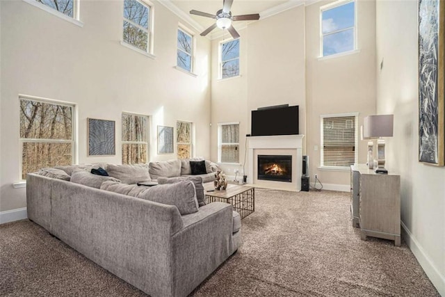 carpeted living room featuring a warm lit fireplace, ceiling fan, ornamental molding, and baseboards