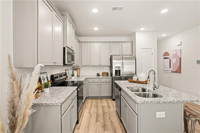 kitchen with sink, a center island with sink, appliances with stainless steel finishes, light stone countertops, and light hardwood / wood-style floors