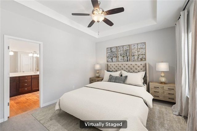 bedroom with ceiling fan, a tray ceiling, light hardwood / wood-style floors, and ensuite bath