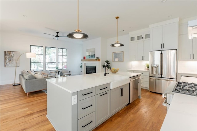 kitchen with appliances with stainless steel finishes, decorative light fixtures, a tiled fireplace, a kitchen island with sink, and light wood-type flooring