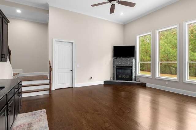 unfurnished living room with dark wood-type flooring, ceiling fan, ornamental molding, and a stone fireplace