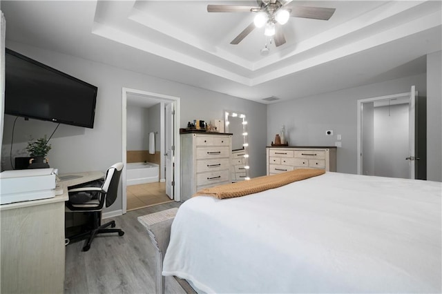 bedroom with light wood-style flooring, an office area, a tray ceiling, connected bathroom, and a ceiling fan