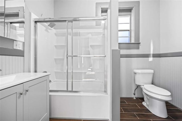 bathroom with wood finish floors, bath / shower combo with glass door, toilet, wainscoting, and vanity