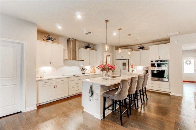 kitchen with wall chimney exhaust hood, a kitchen island with sink, white cabinetry, dark hardwood / wood-style floors, and stainless steel double oven