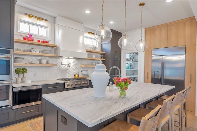 kitchen featuring gray cabinets, an island with sink, custom exhaust hood, built in appliances, and light stone counters