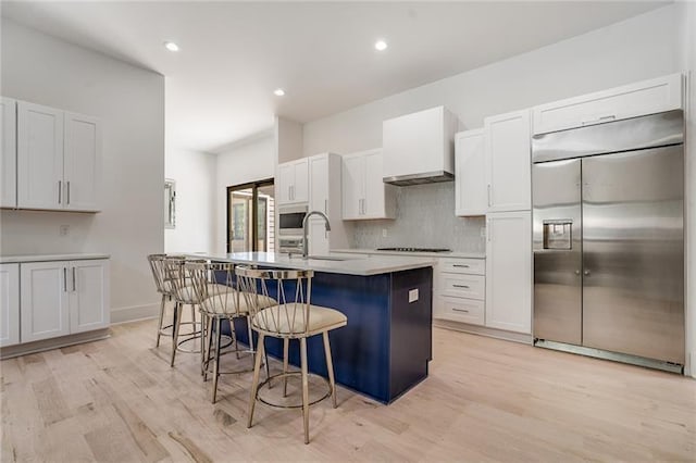 kitchen with light wood-type flooring, wall chimney exhaust hood, stainless steel built in refrigerator, a center island with sink, and a breakfast bar