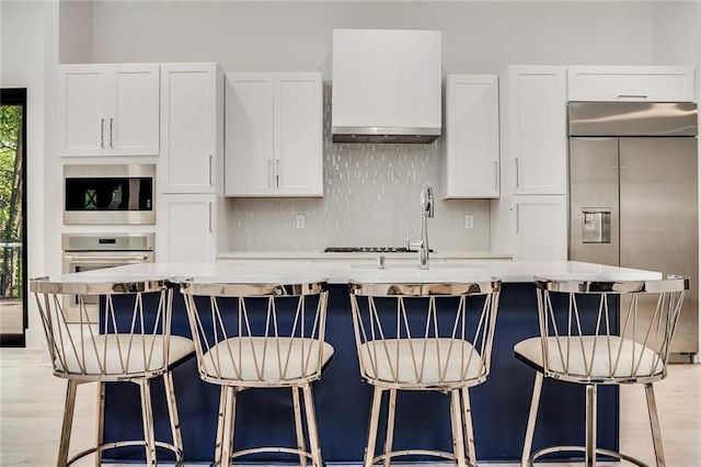 kitchen with white cabinetry, tasteful backsplash, built in appliances, light wood-type flooring, and a center island with sink