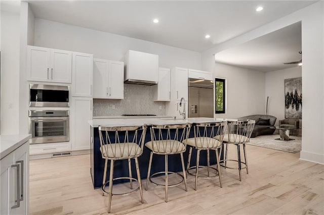 kitchen with built in appliances, light wood-type flooring, a breakfast bar area, backsplash, and a kitchen island with sink