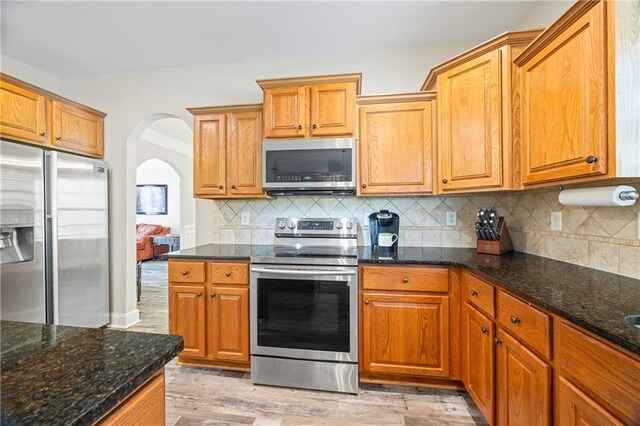 kitchen with appliances with stainless steel finishes, backsplash, light hardwood / wood-style flooring, and dark stone countertops