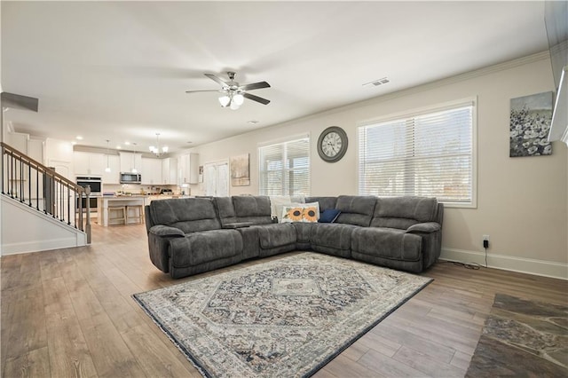 living room with visible vents, baseboards, ornamental molding, ceiling fan with notable chandelier, and light wood-style floors