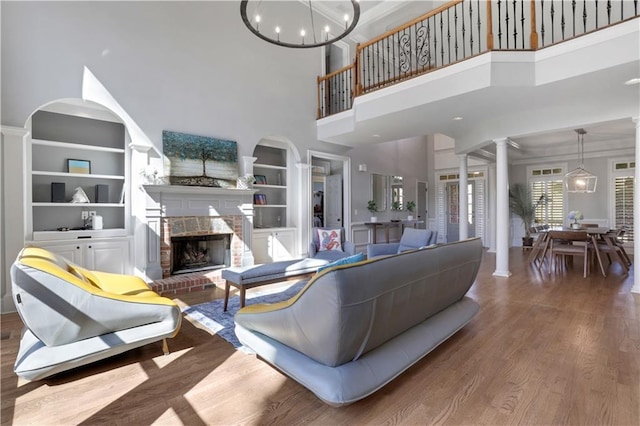 living room featuring built in shelves, wood finished floors, an inviting chandelier, a brick fireplace, and ornate columns