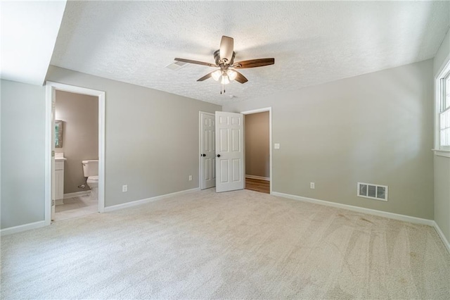 unfurnished bedroom with a textured ceiling, light carpet, baseboards, ensuite bath, and a ceiling fan