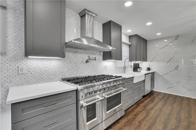 kitchen with wall chimney range hood, dark wood-type flooring, gray cabinetry, backsplash, and stainless steel appliances