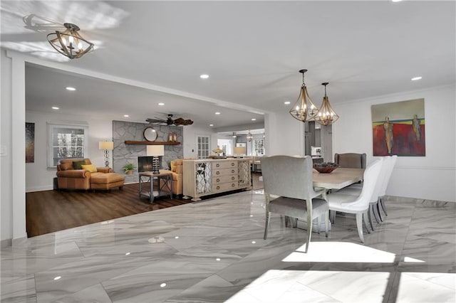 dining space featuring a stone fireplace, ceiling fan with notable chandelier, and ornamental molding