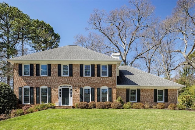 view of front of property with a shingled roof, a front yard, brick siding, and a chimney