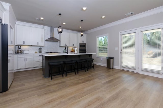 kitchen featuring white cabinetry, wall chimney range hood, decorative light fixtures, a kitchen island with sink, and appliances with stainless steel finishes