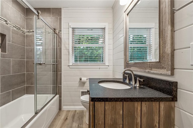 full bathroom with toilet, hardwood / wood-style floors, bath / shower combo with glass door, vanity, and wood walls