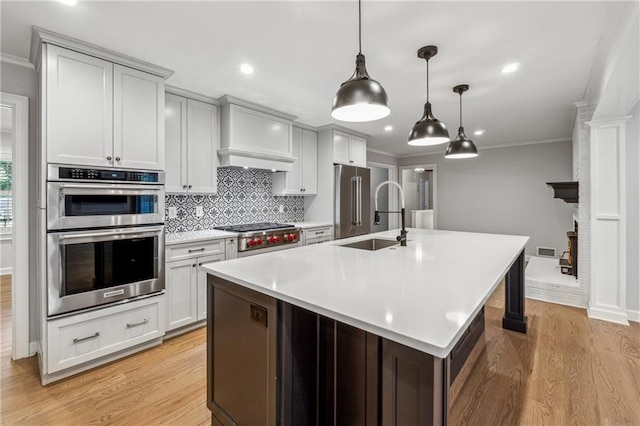 kitchen featuring light wood-type flooring, stainless steel appliances, decorative light fixtures, ornamental molding, and a kitchen island with sink