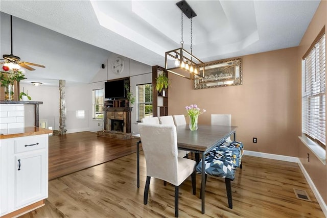 dining room featuring baseboards, light wood finished floors, a fireplace, ceiling fan, and a tray ceiling