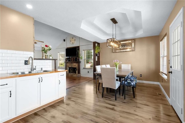 dining room featuring a chandelier, light wood-type flooring, a stone fireplace, baseboards, and a raised ceiling