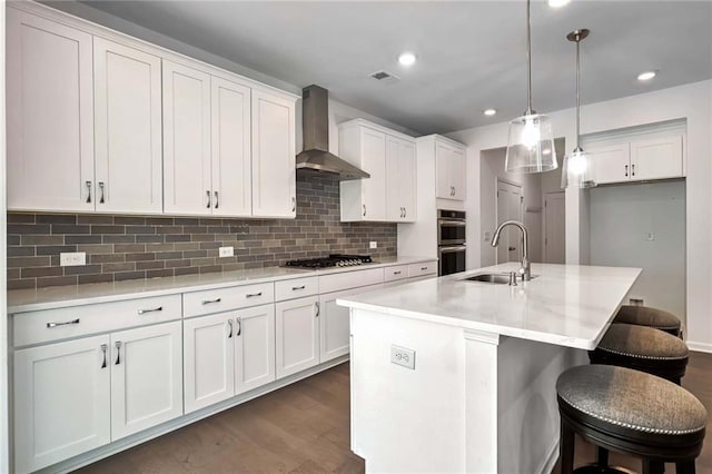 kitchen featuring wall chimney range hood, white cabinets, a center island with sink, decorative light fixtures, and sink